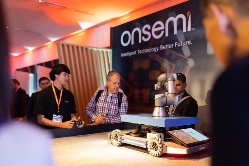An onsemi employee stands next to a robot, explaining something to visitors at a conference.